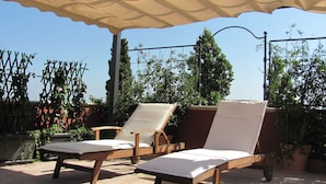 Un área de estar al aire libre techada con dos sillones y una mesa, rodeada de macetas con plantas y un cielo azul despejado.