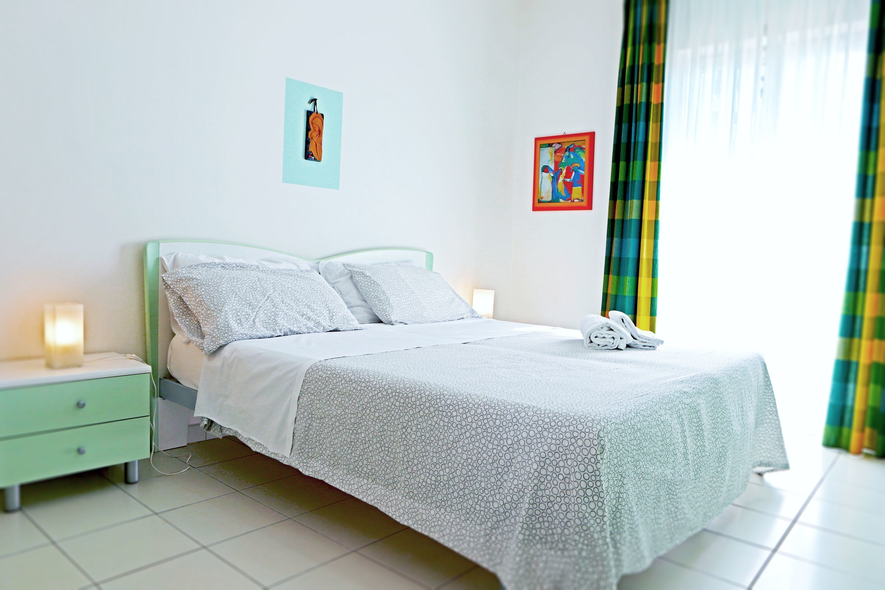 A neatly made bed with a white and gray patterned bedspread, a green nightstand with a lamp, and two wall art pieces.