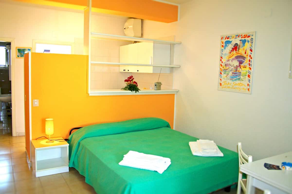 Ein Zimmer mit einem grünen Bett, einem weißen Nachttisch mit Lampe, einem kleinen Regal mit Pflanze und einem Poster an der Wand.