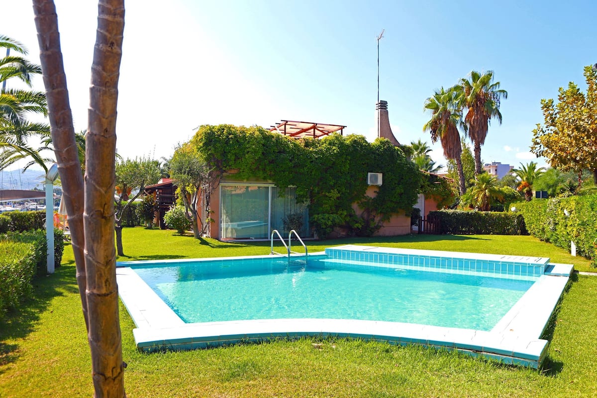 Ein rechteckiger Swimmingpool mit weißem Rand, umgeben von gepflegtem Rasen und Palmen.