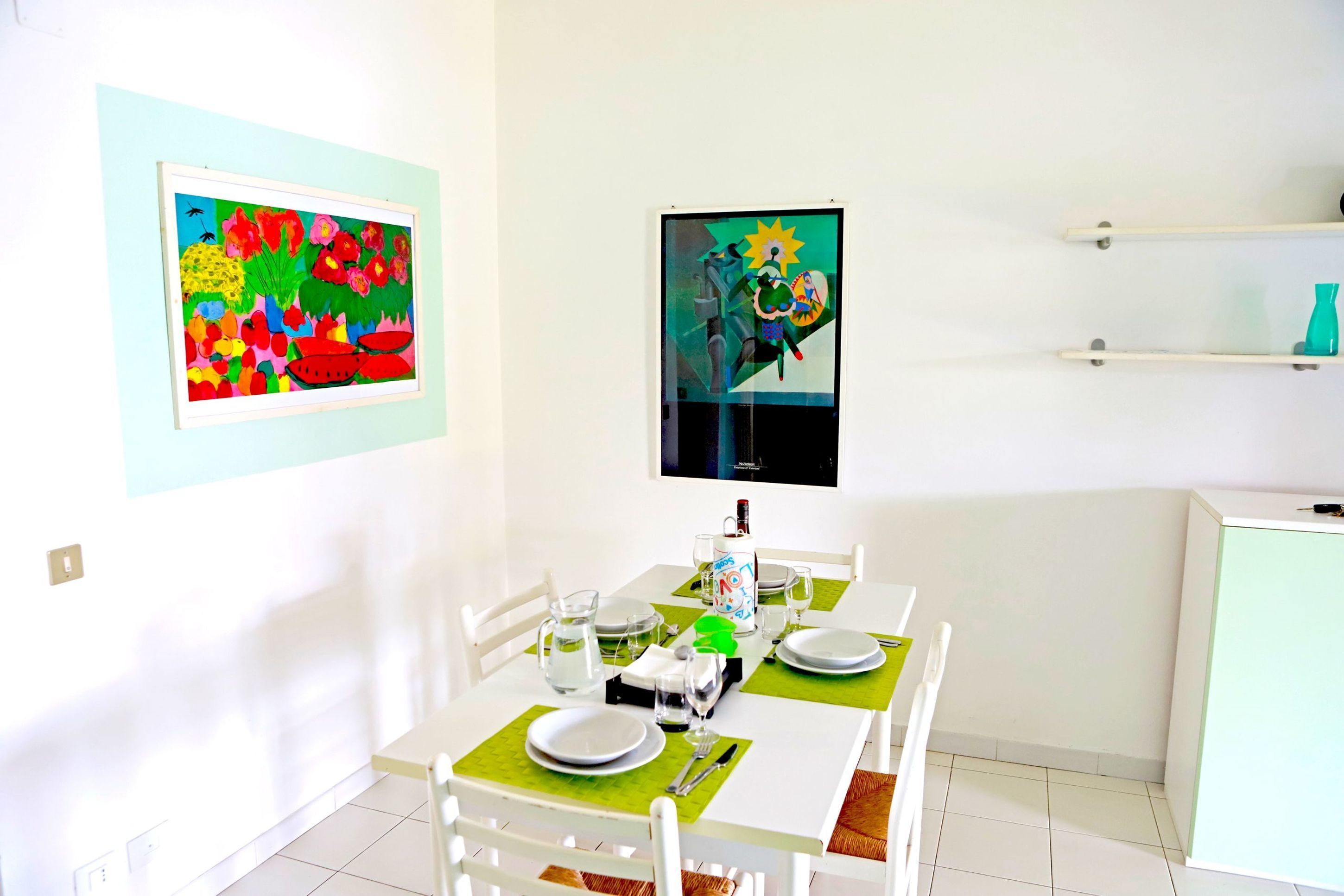 A dining area with a white table set for four, green placemats, white plates, and clear glassware. Two colorful paintings are hung on the walls.