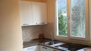 A kitchen with white cabinets, a double sink, and a window with a view of trees. - Camping Valle Romantica (Cannobio)