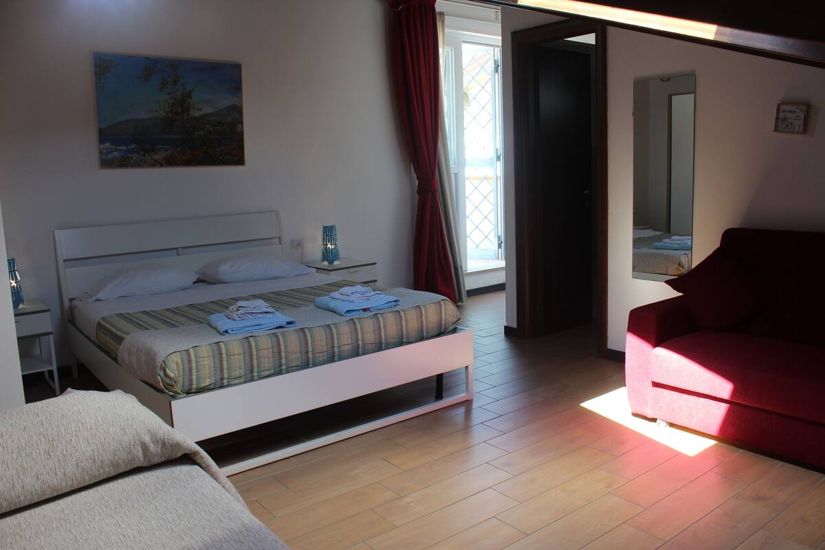 A bedroom with a bed, a red chair, a nightstand, a bedside table, and a window with curtains.