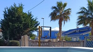 Una piscina cristalina con cubierta de madera al frente, rodeada de palmeras y un edificio azul con techo azul en el fondo.