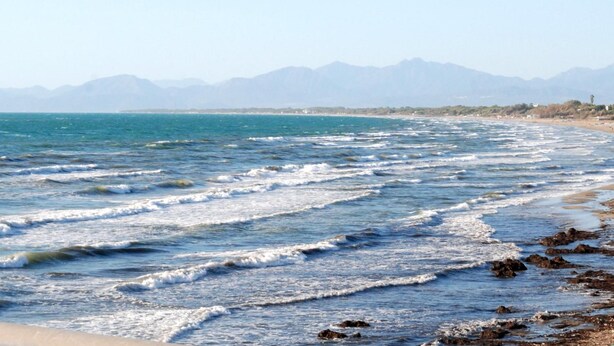 Una playa con olas, costa rocosa y montañas al fondo.
