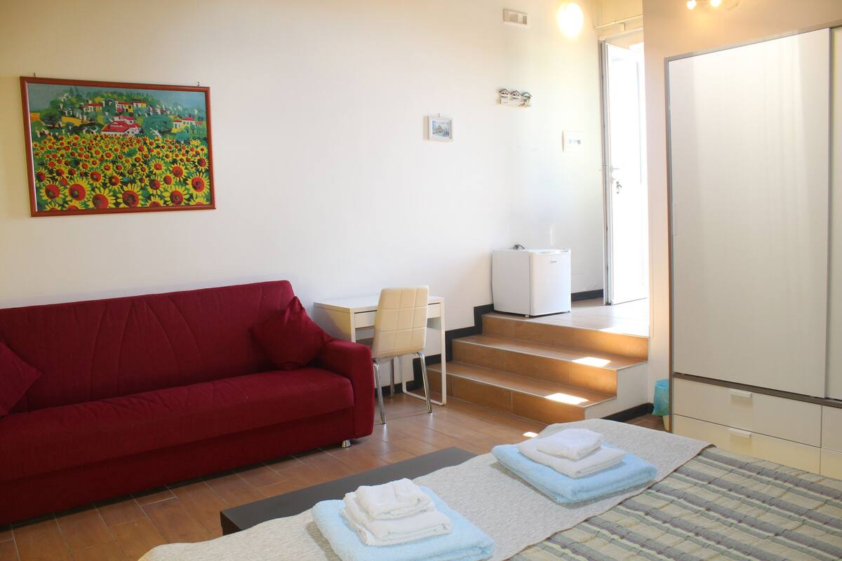 A hotel room with a red sofa, a desk, a chair, and a painting of a sunflower field on the wall.
