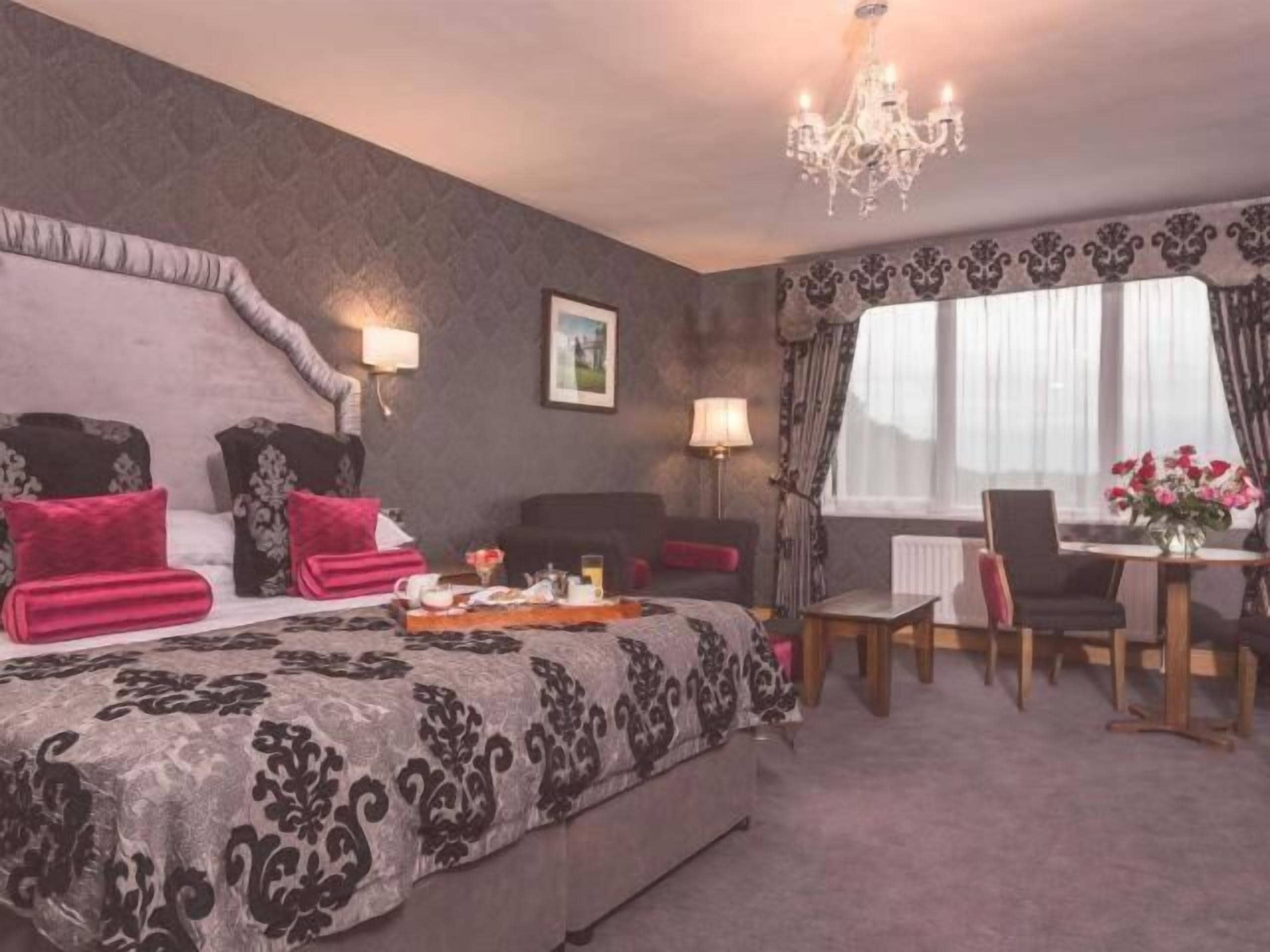A hotel room with a large bed, a chandelier, a dining area with a table and chairs, and a window with curtains.