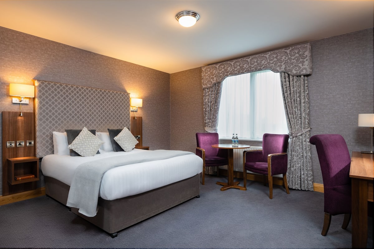 A hotel room with a bed, two chairs, a small table, and a window with curtains.