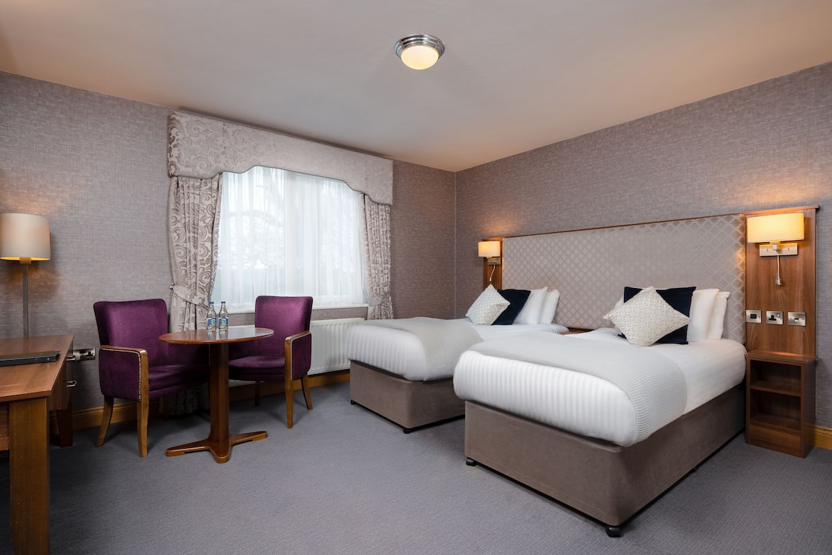 A hotel room with two beds, a small table, purple chairs, and a window with curtains.