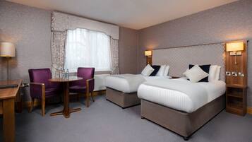 A hotel room with two beds, a small table, purple chairs, and a window with curtains.