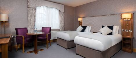 A hotel room with two beds, a small table, purple chairs, and a window with curtains.