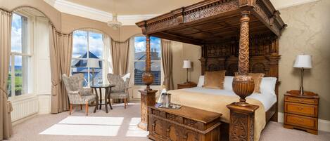 A four-poster bed with a wooden headboard, a sitting area with a table and chairs, a bedside table with a lamp, and a nightstand with drawers.
