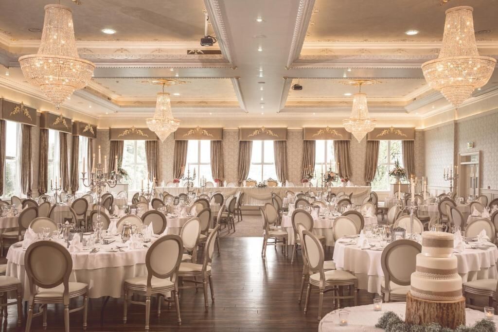 A banquet hall with round tables, chandeliers, and a tiered cake centerpiece.