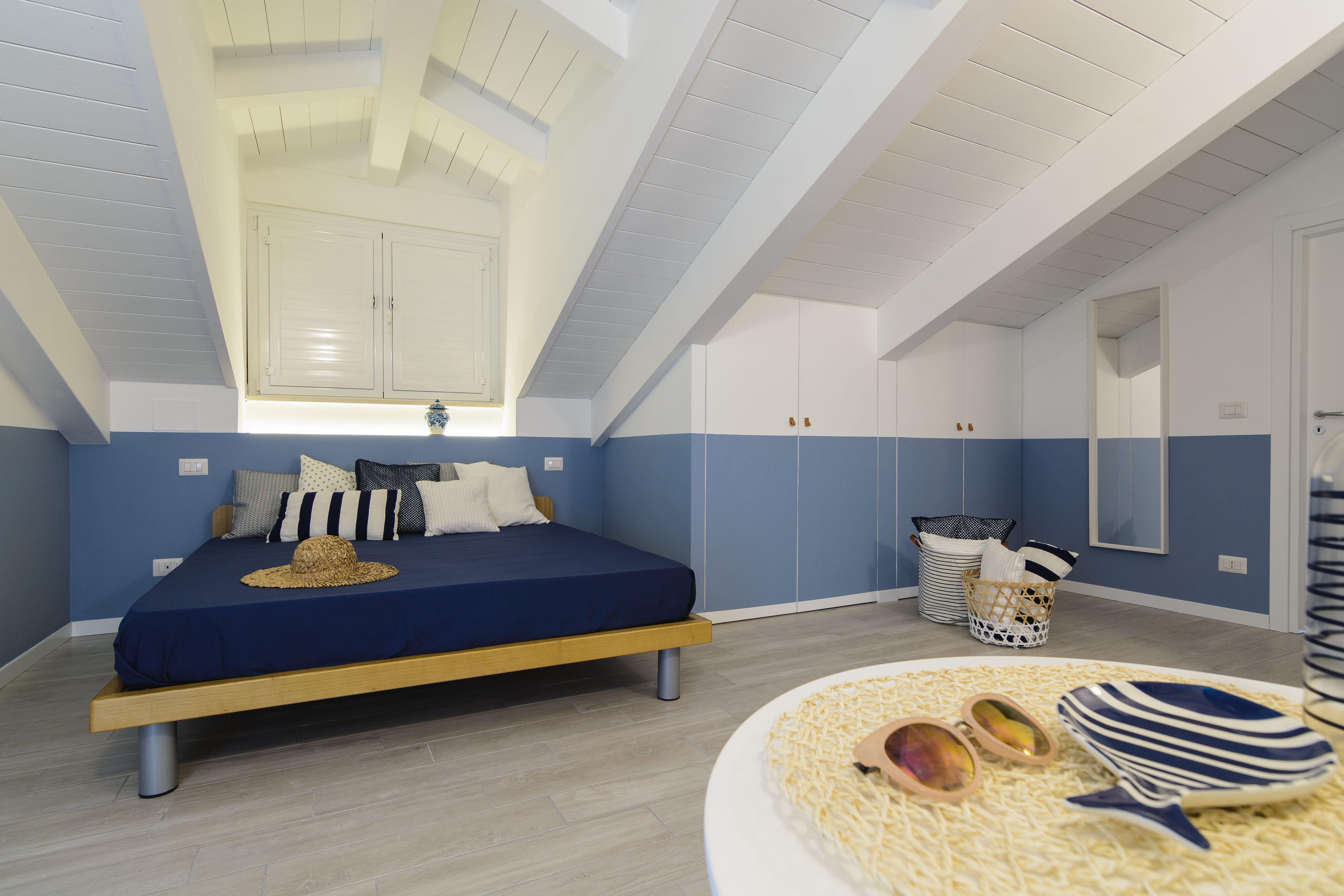 A bedroom with a bed, a window, a mirror, and a small table with a hat and sunglasses.