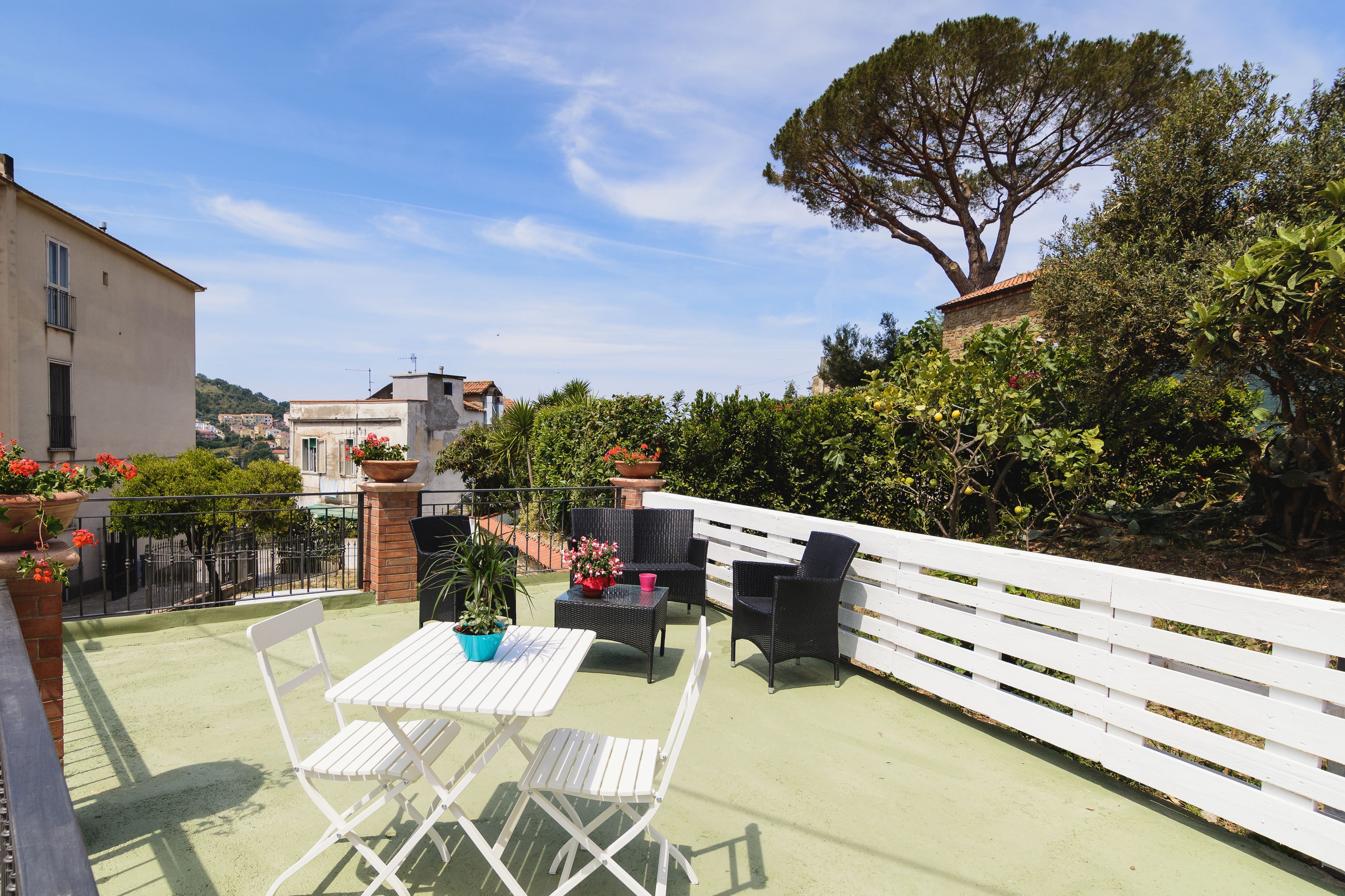 A rooftop patio with a white table and chairs, surrounded by a white fence, and overlooking a cityscape.