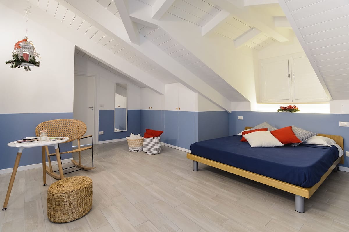 A modern bedroom with a sloped ceiling, a bed with multiple pillows, a wicker chair, and a small table.