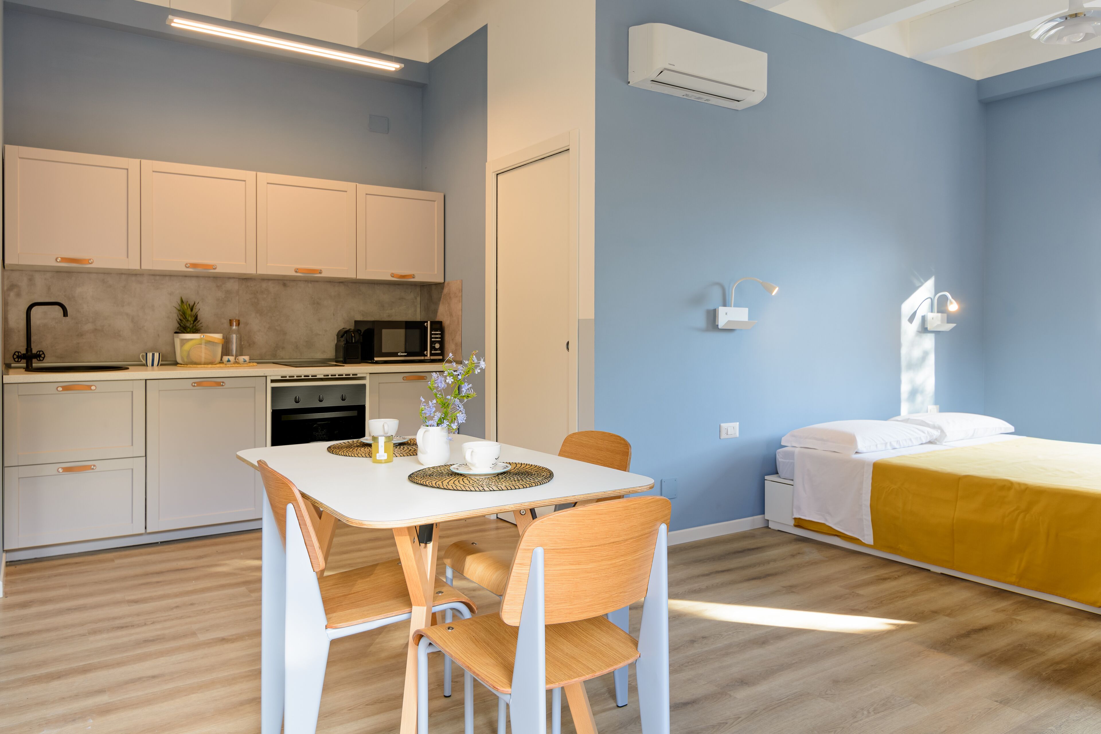 Cocina pequeña con gabinetes blancos, microondas y pileta. Hay una mesa de comedor con sillas y una cama con cubrecama amarillo.