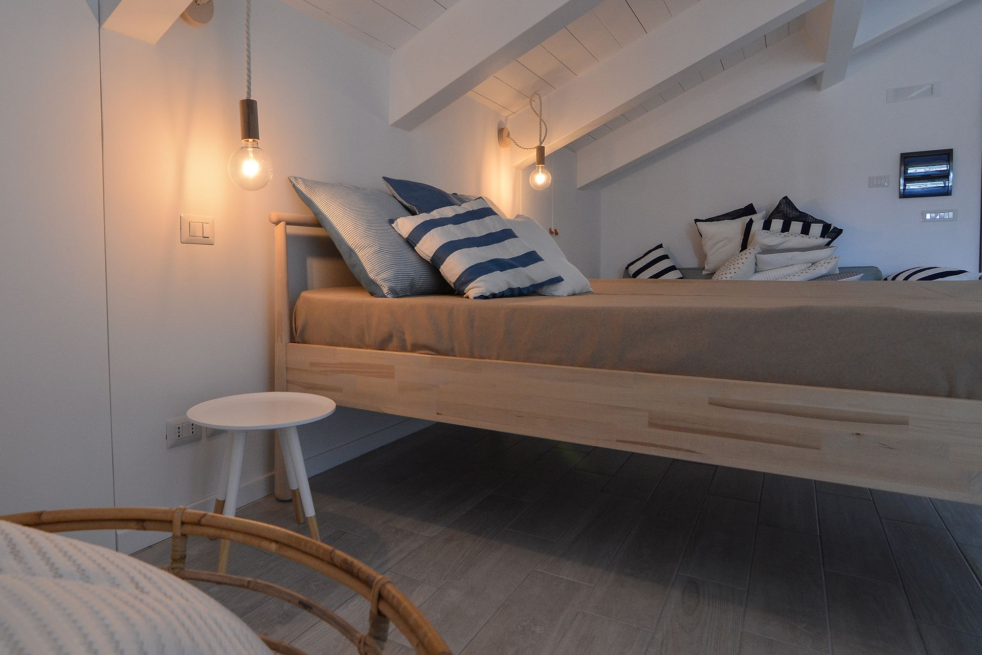 Un dormitorio estilo loft con una cama de madera, dos bombillas colgantes, una mesita redonda y una silla de mimbre.