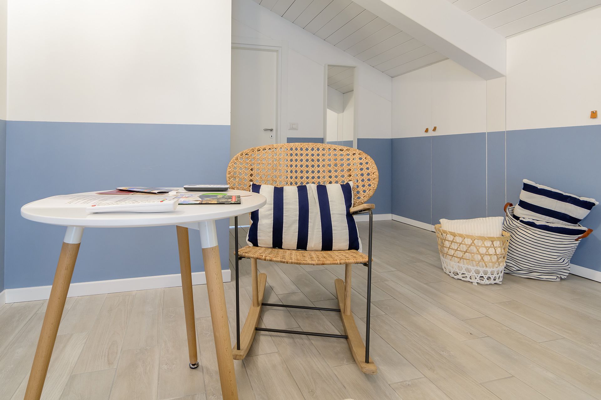 A room with a wicker chair, a small round table, and a striped cushion. There is a wicker basket and a striped pillow on the floor.