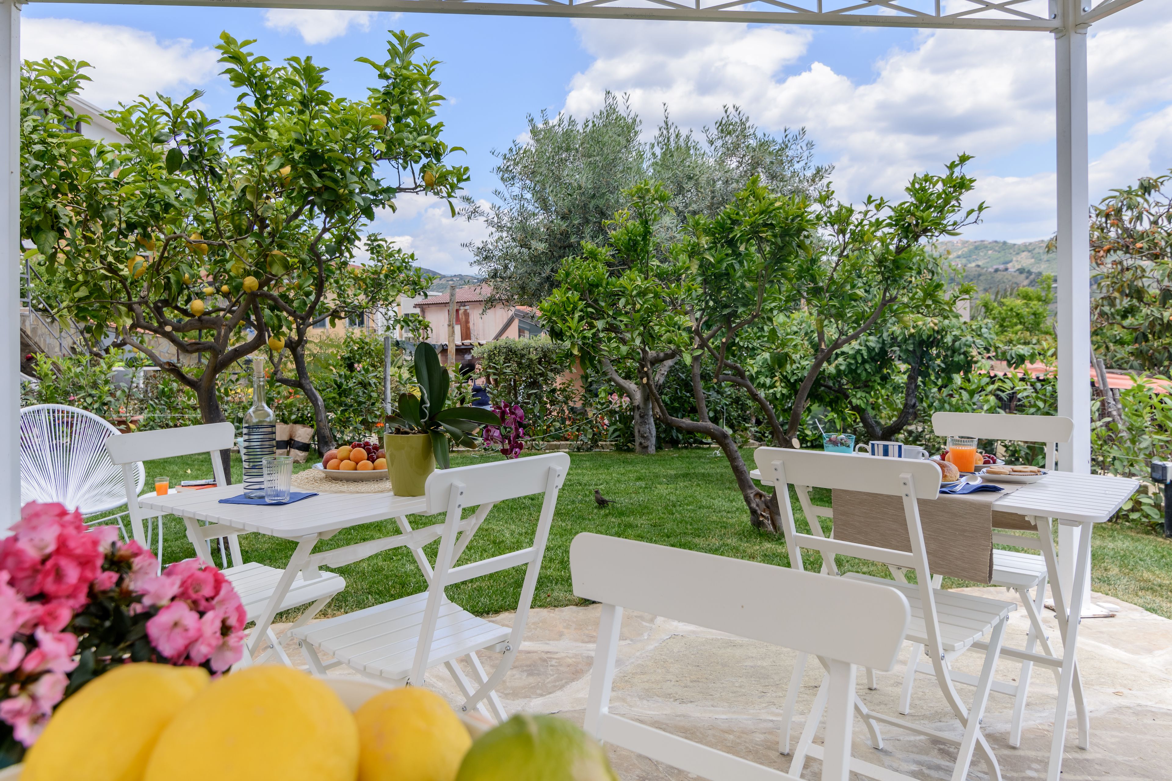 Un patio al aire libre con mobiliario blanco, una mesa con frutero y una planta en maceta.