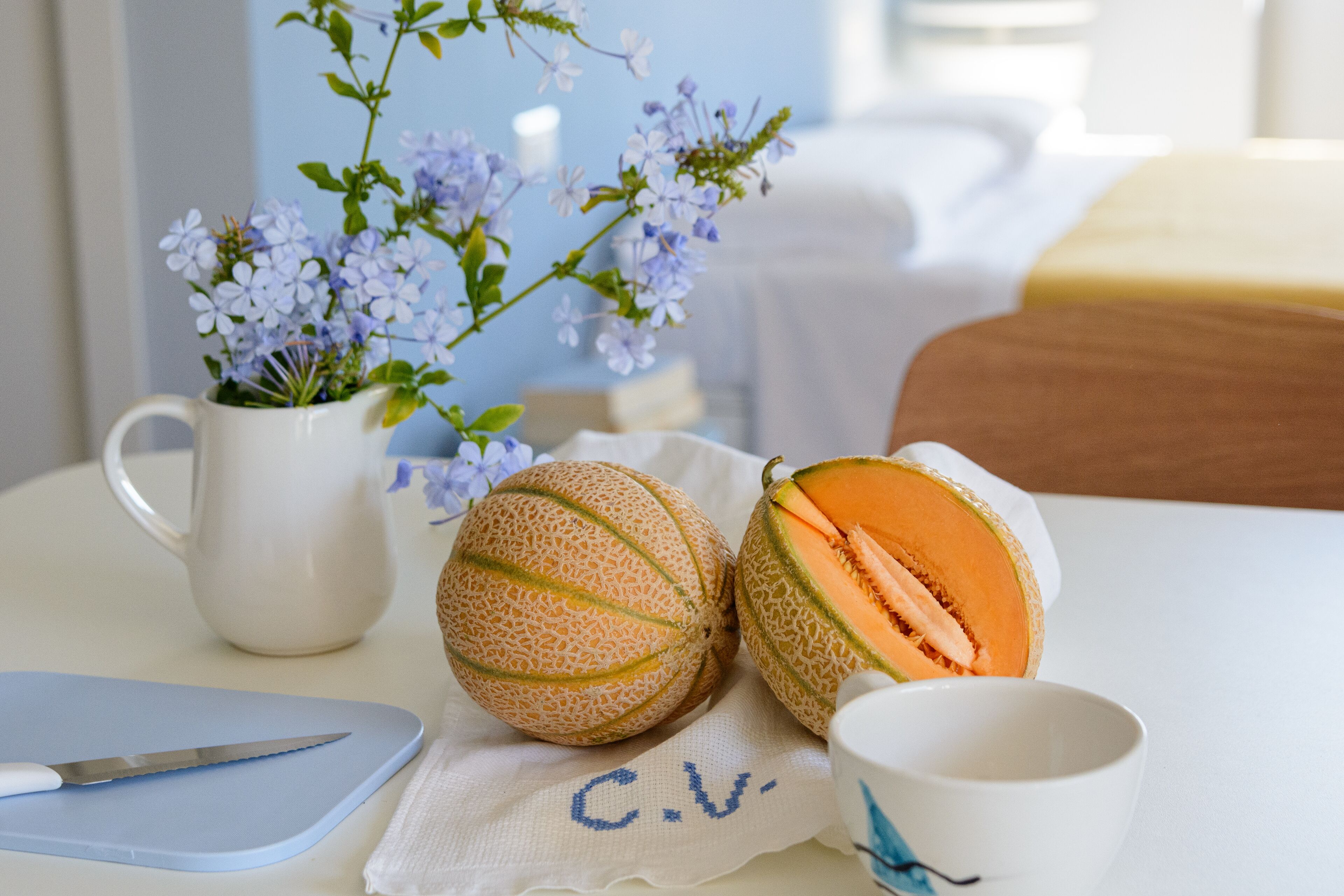 A table with a white pitcher of flowers, a halved cantaloupe, a blue cutting board, and a knife.