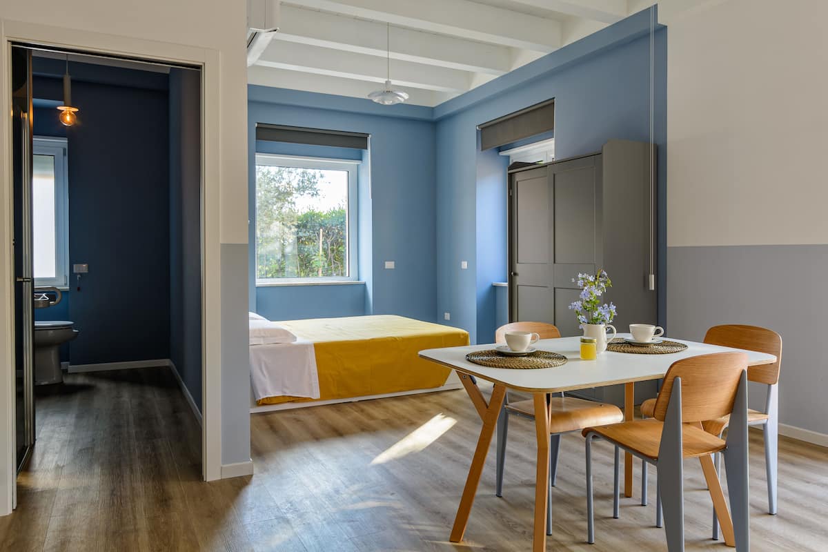 A room with a bed, a dining table with chairs, a kitchen area, and a window with a view of greenery.