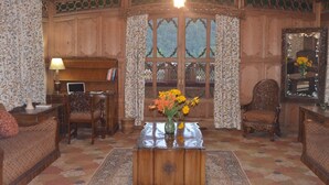 A traditional room with a wooden paneled wall, a large window with curtains, a wooden coffee table with a vase of flowers, and two upholstered chairs. - New Alexandra Houseboats (Srinagar)