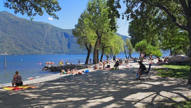 Una playa junto al lago con gente tomando el sol, nadando y paseando en bote.