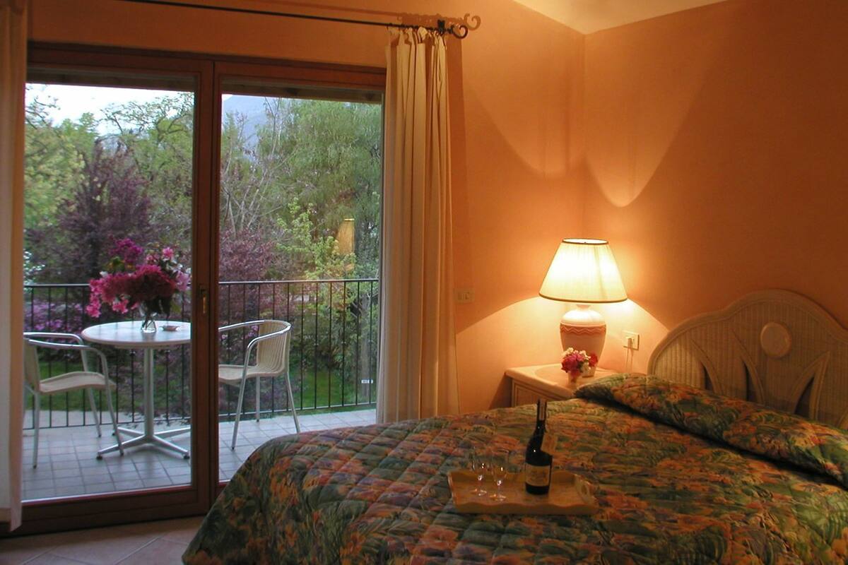 A bedroom with a bed, a bedside table with a lamp, a window with curtains, and a balcony with a table and chairs.