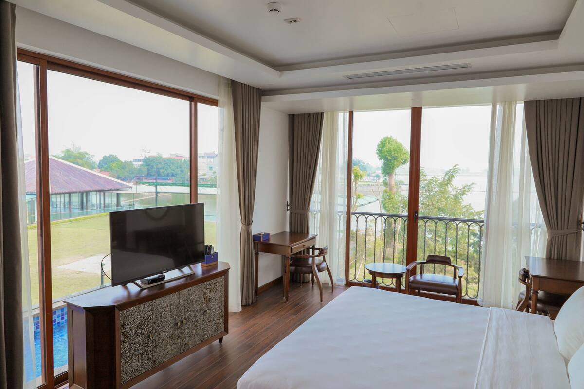 A hotel room with a large bed, a TV on a wooden cabinet, a desk with a chair, and a balcony with a view of a tennis court and buildings.