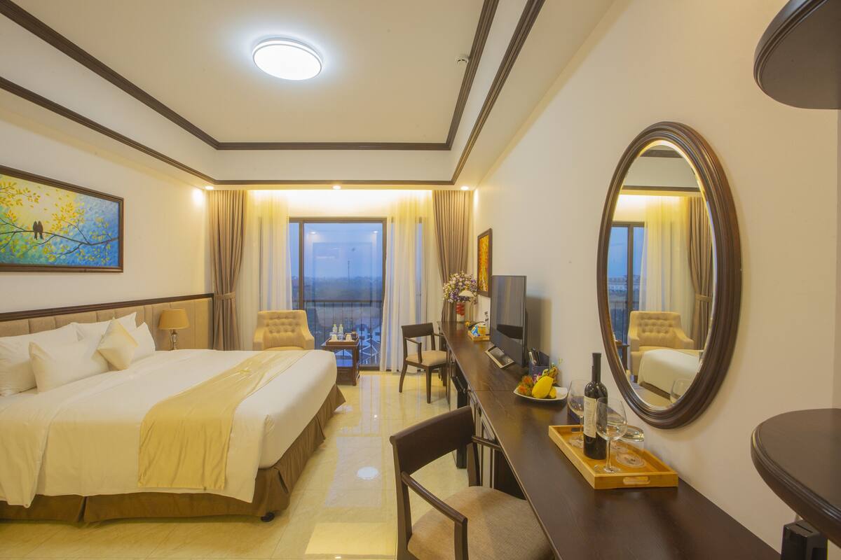A hotel room with a large bed, a desk with a mirror, a TV, and a balcony with curtains.