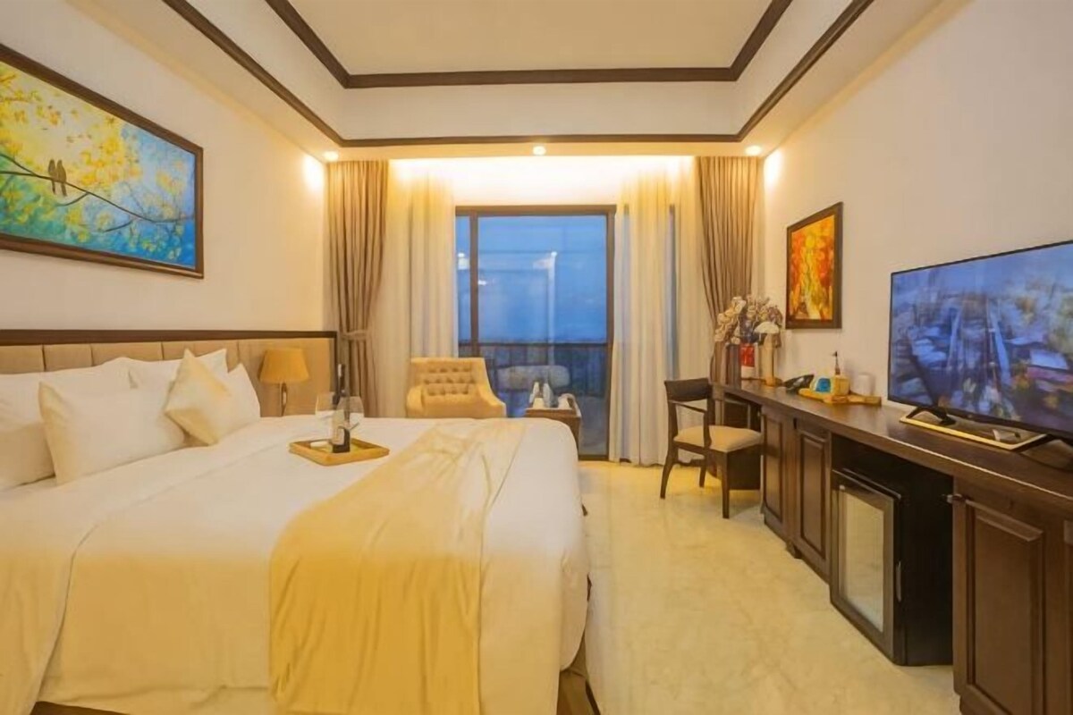 Habitación de hotel con una cama grande, un escritorio con televisor, una silla y un balcón con cortinas.