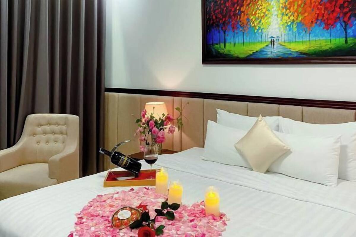 A hotel room with a bed, a chair, a painting, and a table with a bottle, glasses, and flowers.