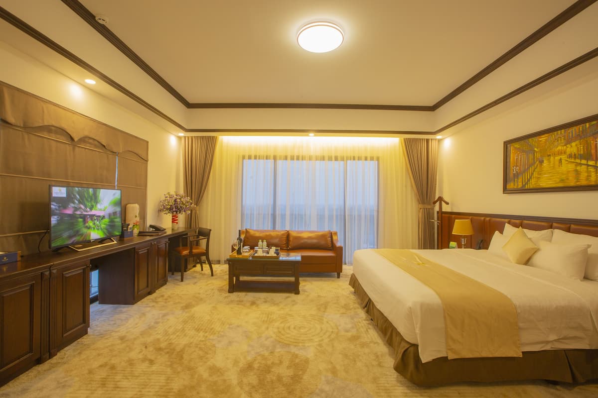A hotel room with a large bed, a desk with a television, a coffee table, and a view of a building through the window.