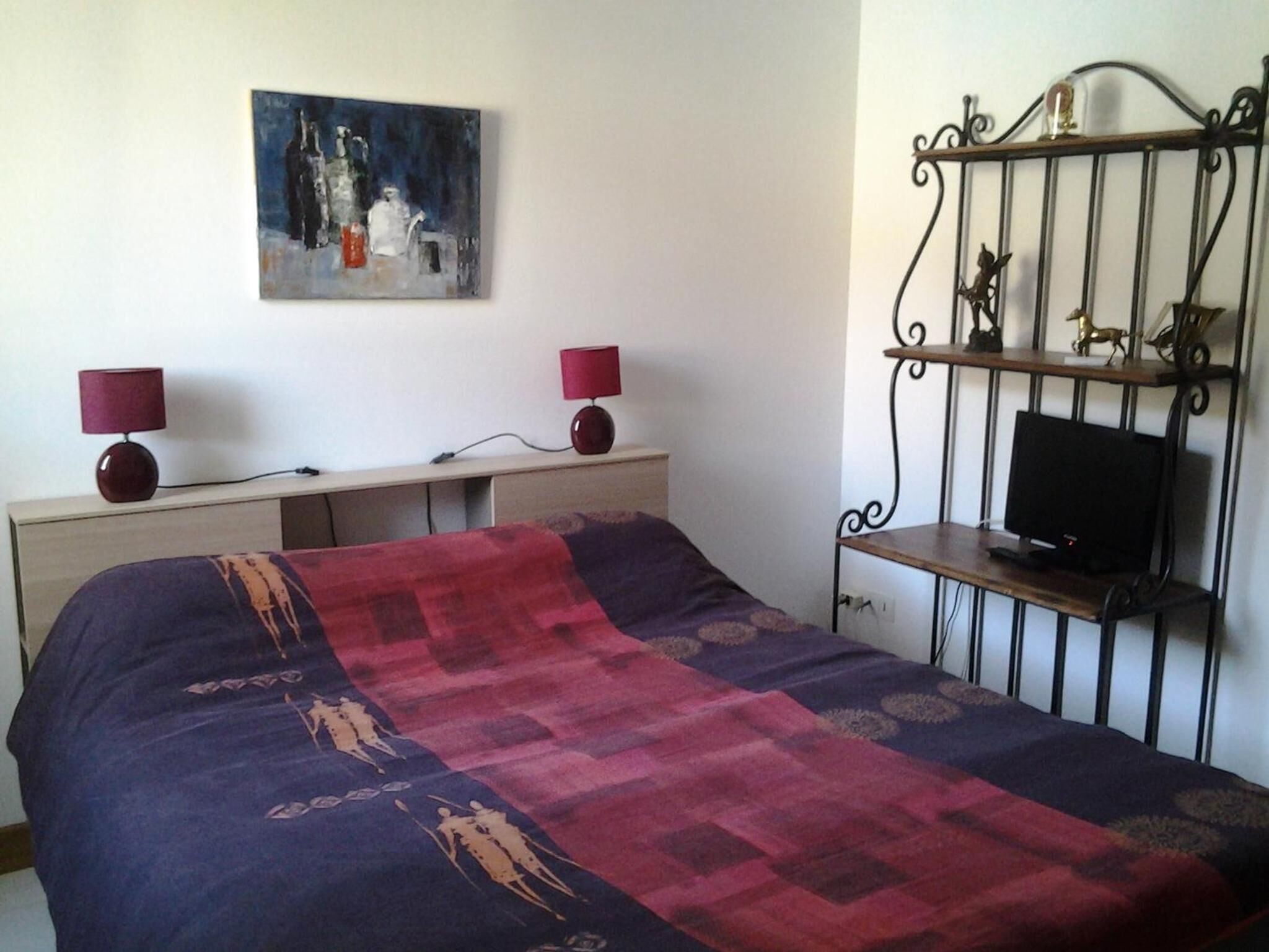 A bedroom with a bed, a desk, a TV, and a painting on the wall.