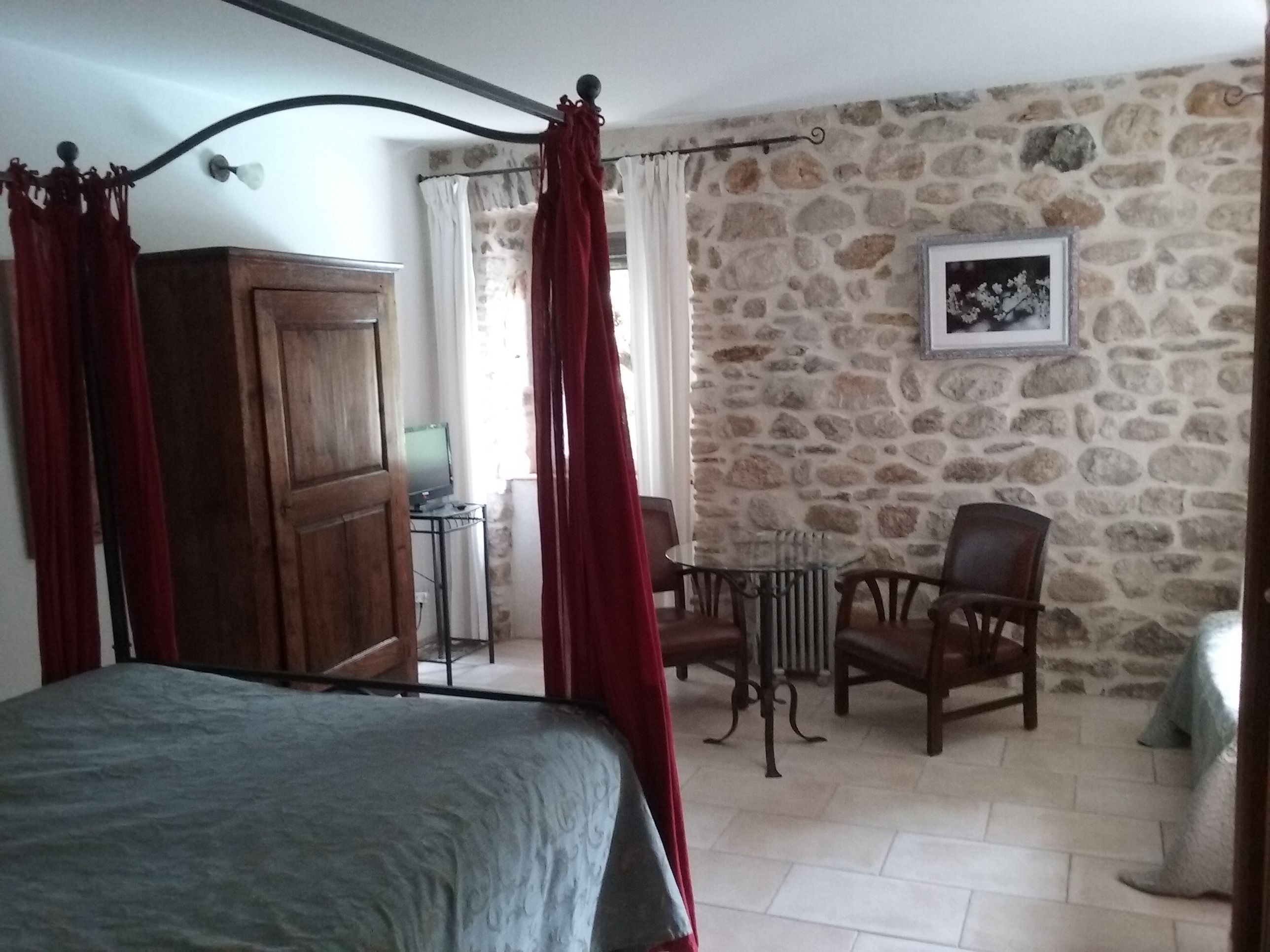 A bedroom with a bed, a wooden wardrobe, a stone wall, a chair, and a table with a lamp.