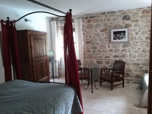 A bedroom with a bed, a wooden wardrobe, a stone wall, a chair, and a table with a lamp.