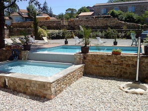 A pool area with two rectangular pools, surrounded by stone walls and pebbles. There are lounge chairs and potted plants around the pools.