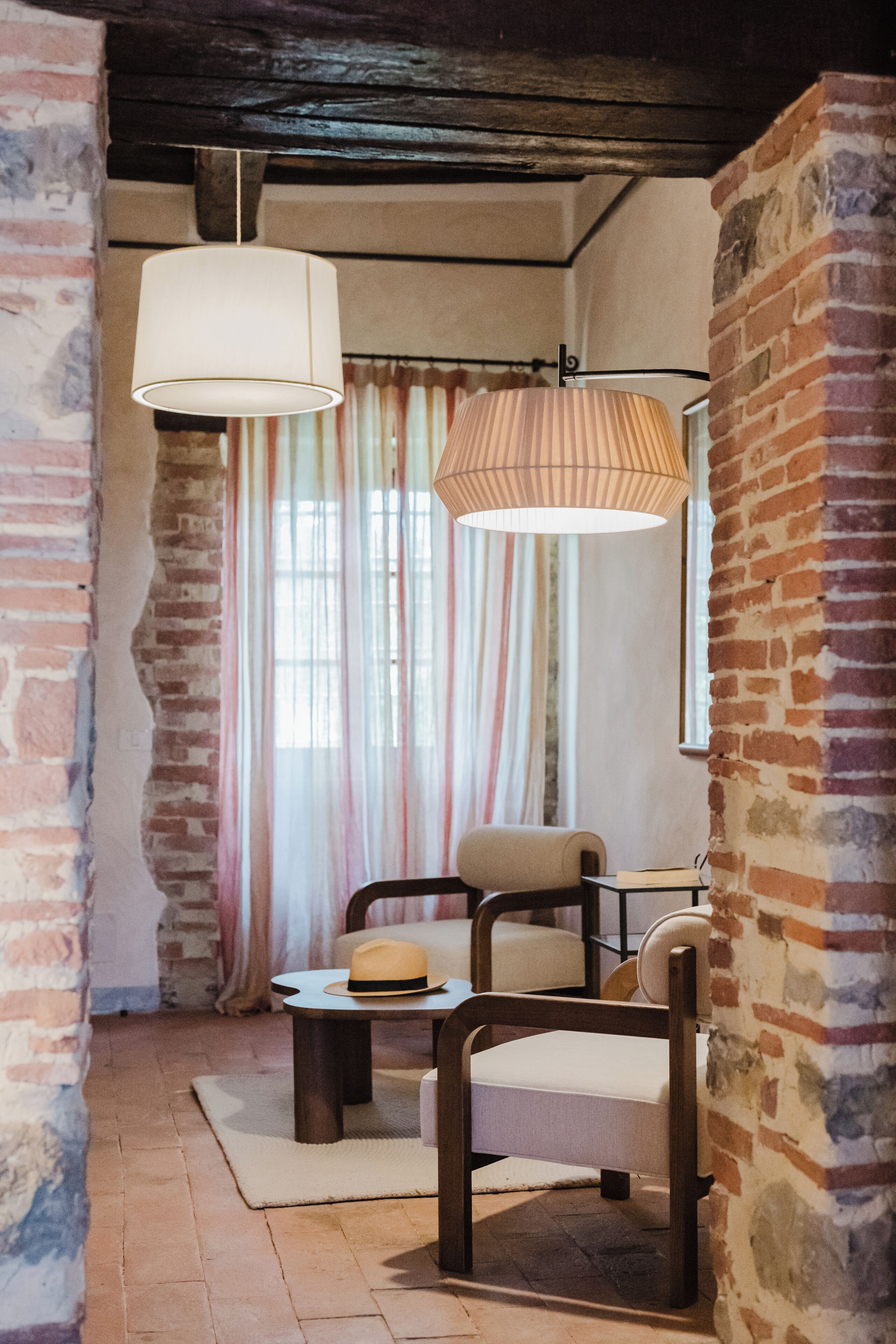 A room with exposed brick walls, a wooden ceiling, a large window with curtains, a chair, a table, and a hanging lamp.