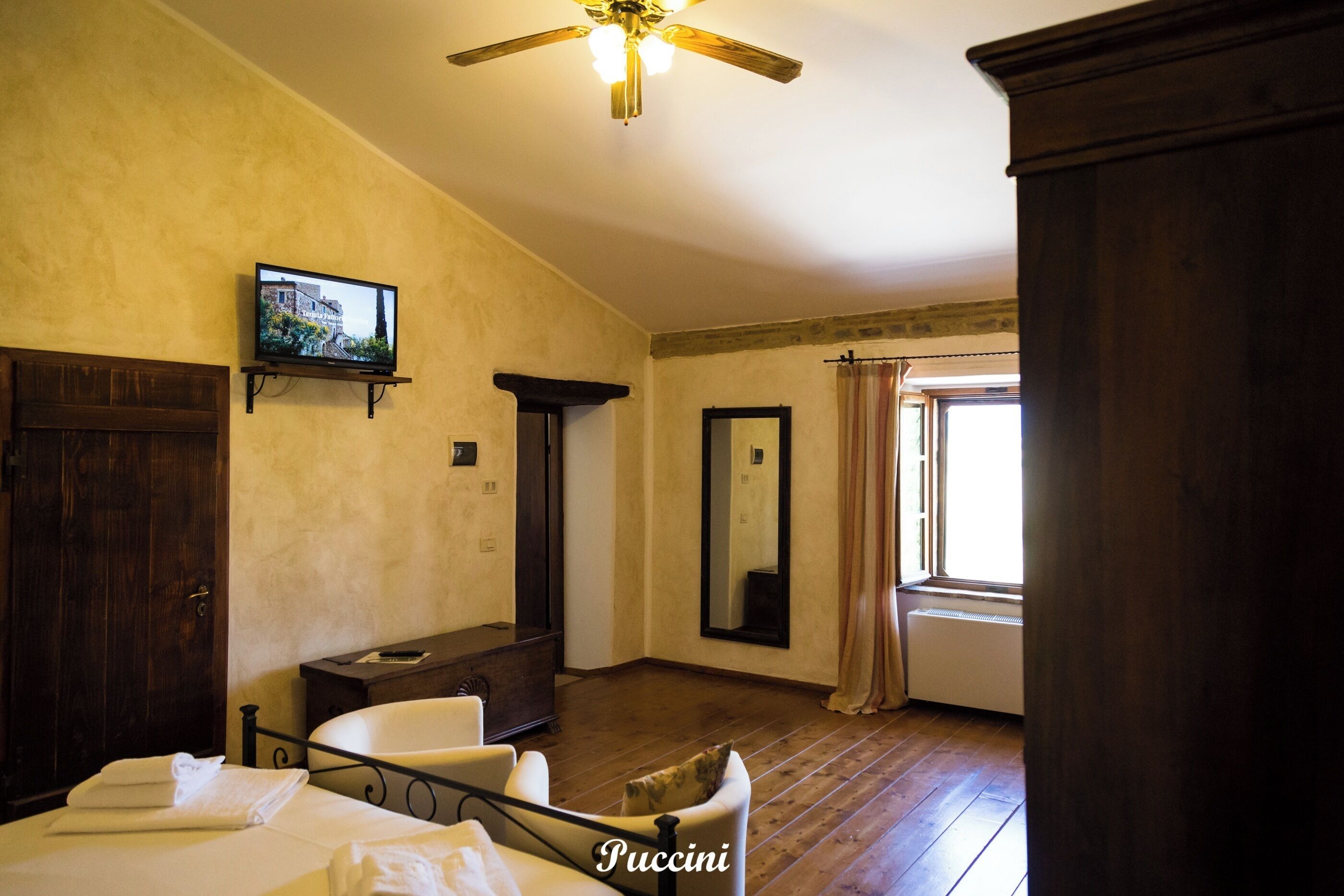 Habitación de hotel con cama, una silla, televisor, ventilador de techo, ventana con cortinas y un armario de madera.