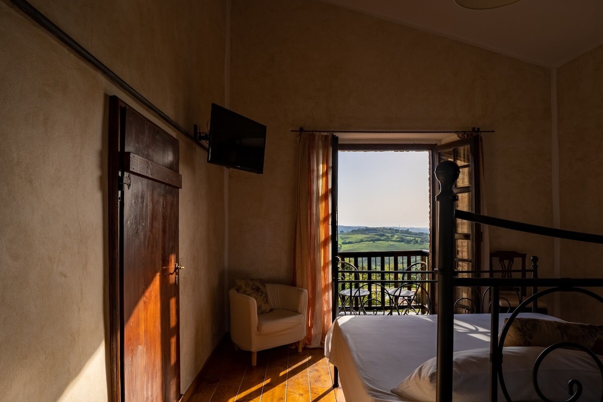 A bedroom with a bed, a chair, a TV mounted on the wall, and a balcony with a view of the countryside.