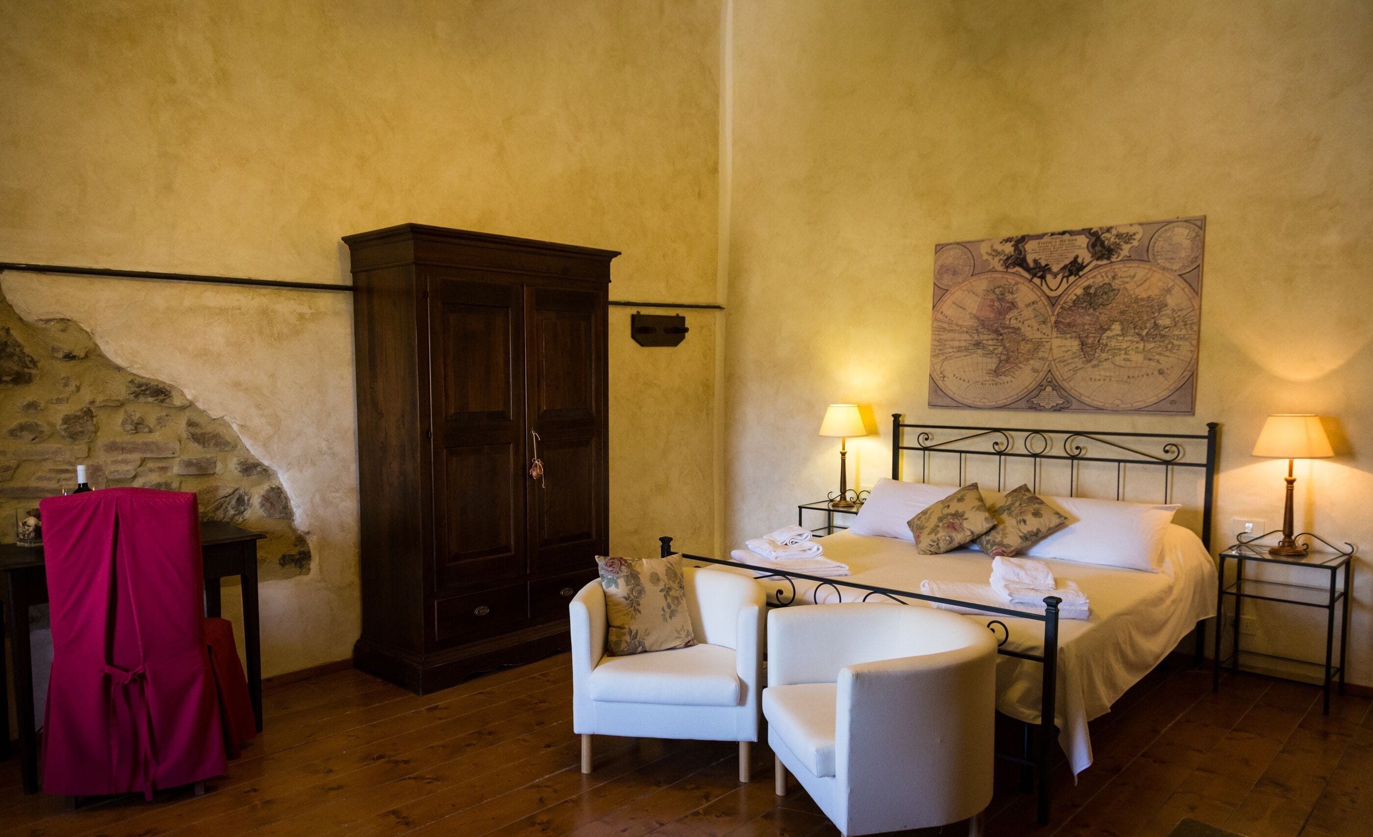 A bedroom with a bed, two bedside tables with lamps, a wooden wardrobe, a chair, and a map on the wall.
