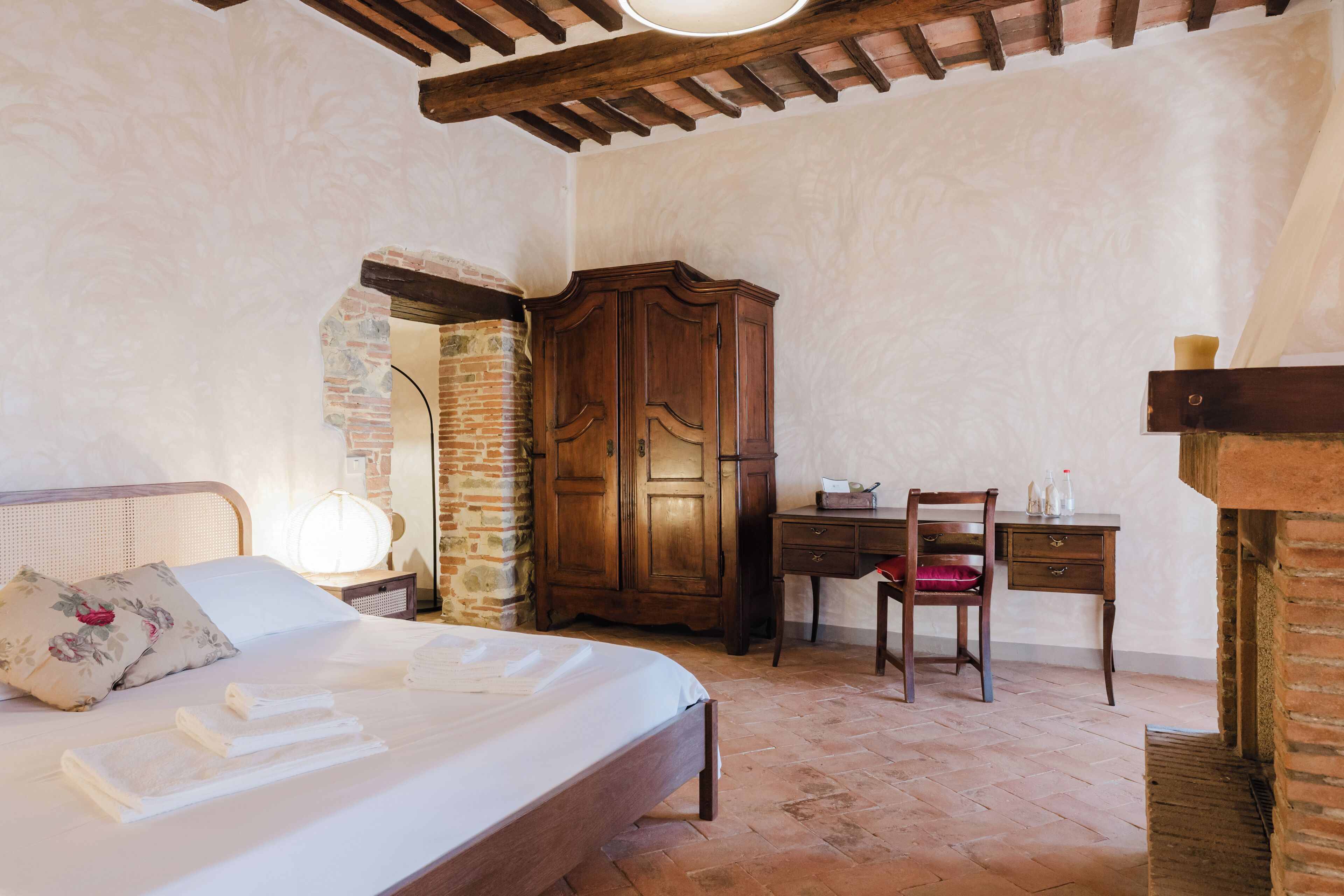 A bedroom with a bed, a wooden wardrobe, a desk with a chair, and a stone wall.