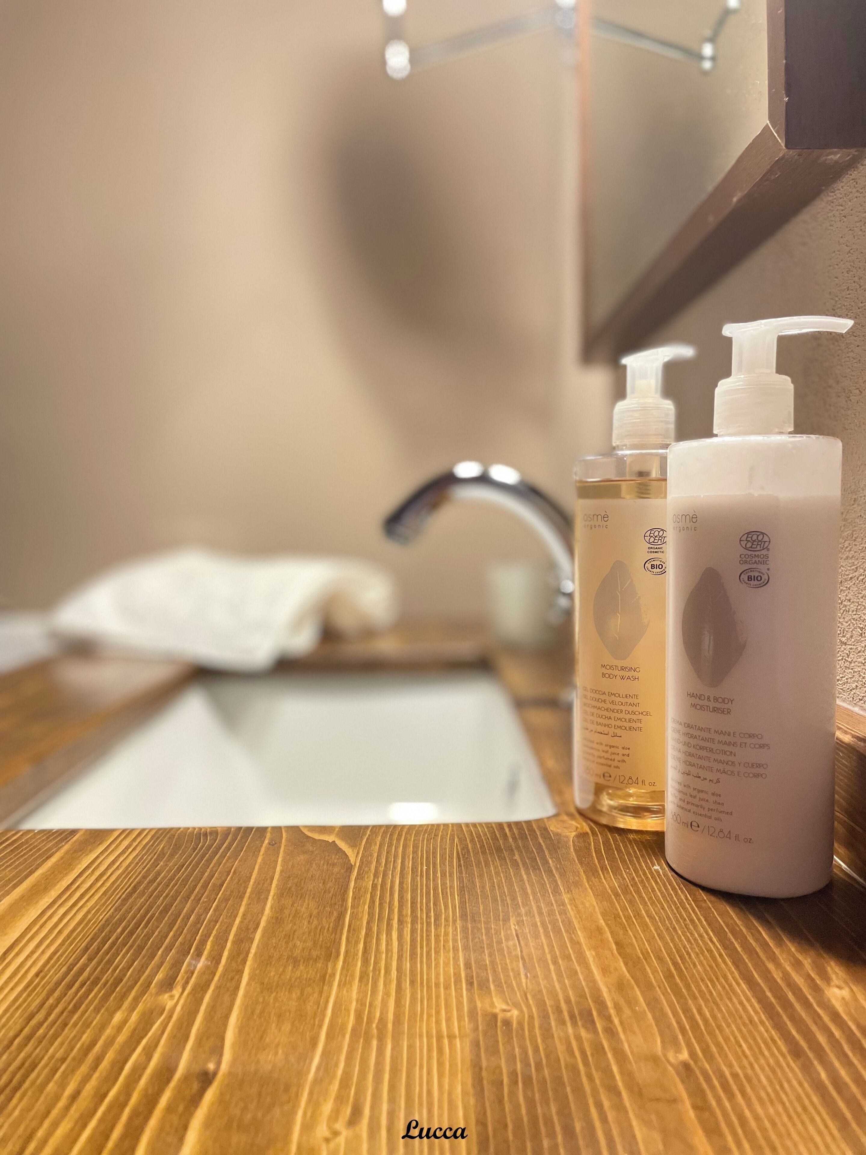 Un piano di lavoro in legno con uno specchio sopra che riflette un lavabo da bagno. Sul lavabo, due flaconi di sapone mani e corpo con l'etichetta 'COSMOS ORGANIC'.