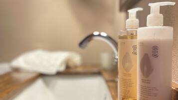 Un piano di lavoro in legno con uno specchio sopra che riflette un lavabo da bagno. Sul lavabo, due flaconi di sapone mani e corpo con l'etichetta 'COSMOS ORGANIC'.