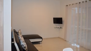 A modern living room with a sofa, a flat-screen TV mounted on the wall, and a large window with sheer curtains. - AZ New Pereybere Beach Apartment (Grand-Baie)