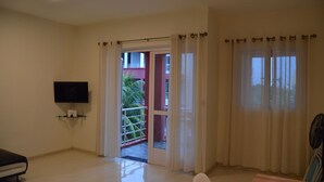 A hotel room with a flat-screen TV on a shelf, a bed, a chair, and a balcony with a view of a building and palm tree. - AZ New Pereybere Beach Apartment (Grand-Baie)