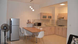 A modern kitchen with a refrigerator, microwave, sink, and wooden cabinets. There is a dining table with chairs and a fan. - AZ New Pereybere Beach Apartment (Grand-Baie)