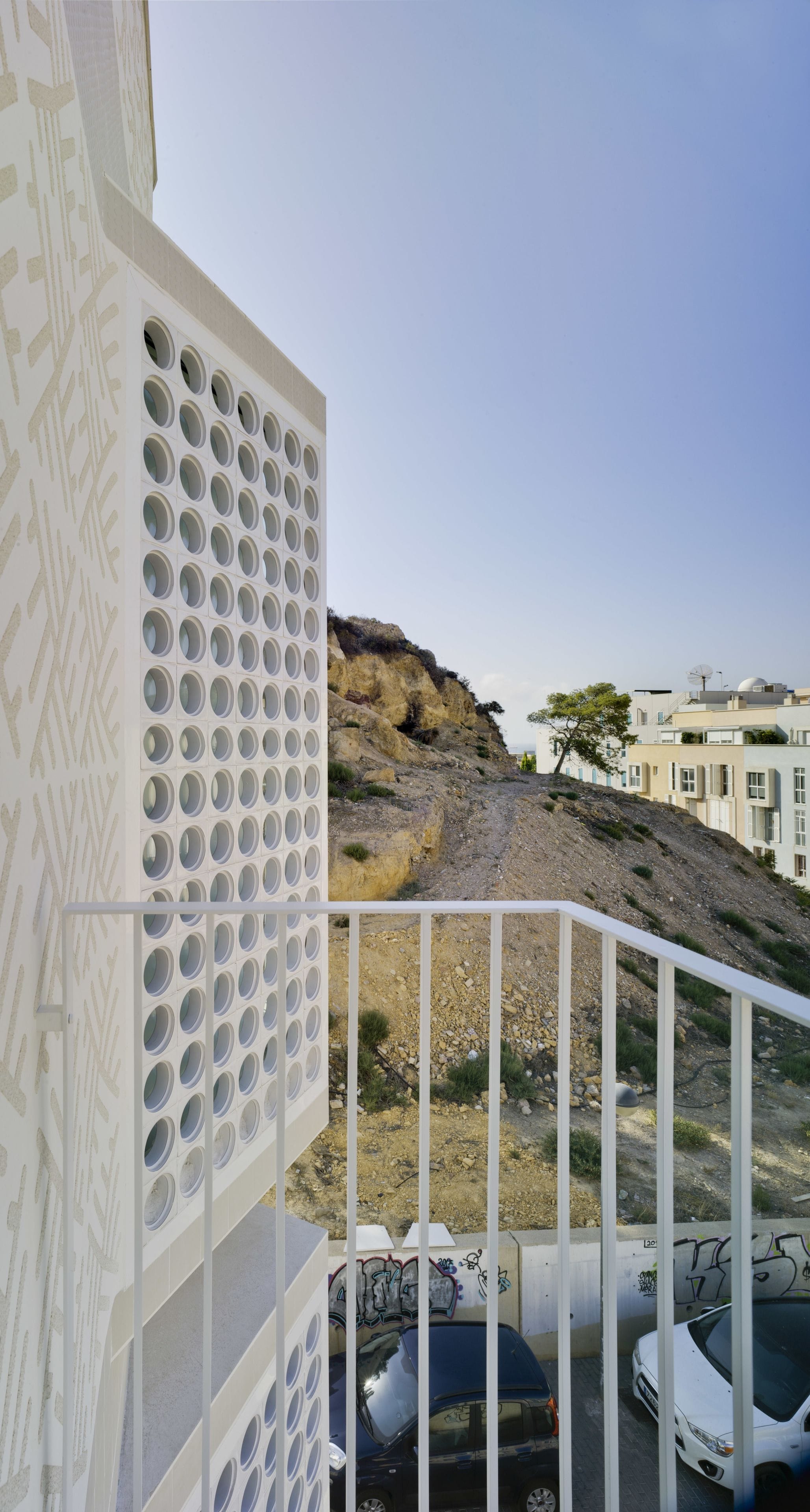 A residential area with a white building featuring circular windows, a rocky hillside, and parked cars.
