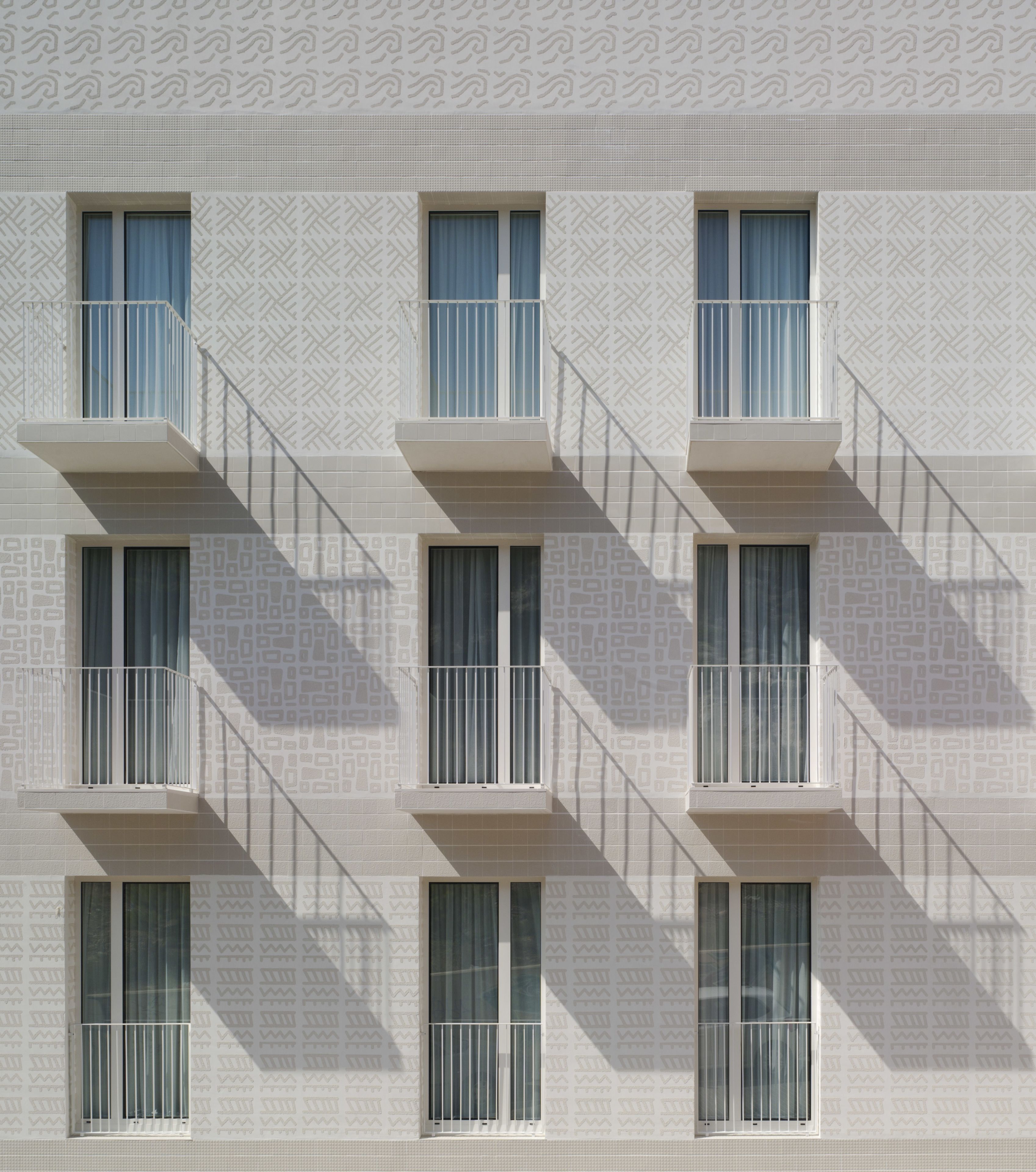 A building facade with a repeating geometric pattern, multiple windows with white railings, and a series of balconies.